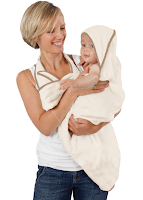 A smiling woman with short blonde hair, wearing a white vest top and jeans, holds a baby wrapped in a cream-coloured hooded towel. The baby looks content, and both face each other, set against a plain white background.