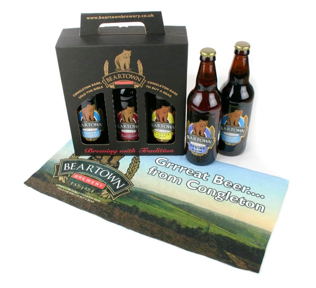 A gift set of Beartown Brewery beers includes four bottles in a black carrier with a bear logo. Three beer bottles stand beside it. In front, a rectangular mat reads "Grreat Beer... from Congleton" with a landscape and brewery logo.