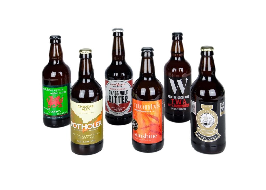 Six different beer bottles stand upright on a white background, each with a distinct label and colour. The labels display various brewery names, beer styles, and colourful designs, highlighting their individual branding and origin.
