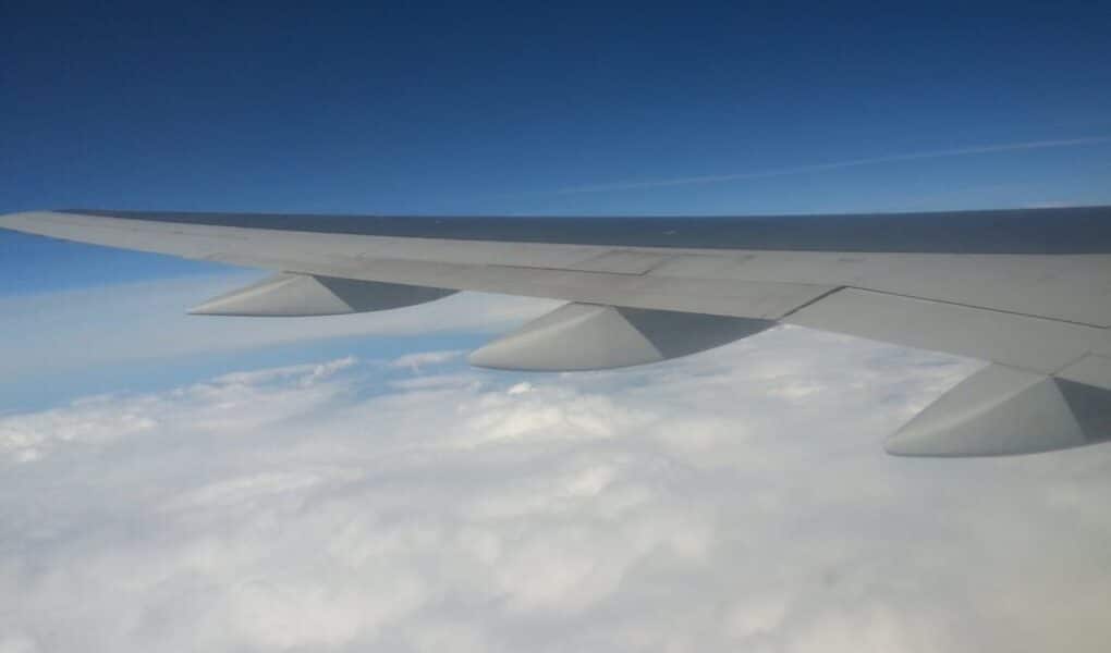 A view from an aeroplane window showing a large grey wing extending over a blanket of white clouds below, with a deep blue sky above. The wing’s flaps and engines are visible, creating a sense of altitude and peaceful flight.
