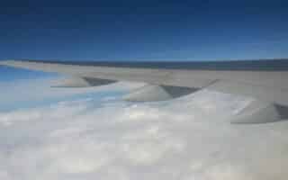 A view from an aeroplane window showing a large grey wing extending over a blanket of white clouds below, with a deep blue sky above. The wing’s flaps and engines are visible, creating a sense of altitude and peaceful flight.