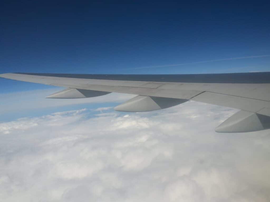 A view from an aeroplane window showing a large grey wing above a thick layer of white clouds, with a clear blue sky overhead. The image captures the scene from inside the aircraft during flight, emphasising the wing and cloudscape below.