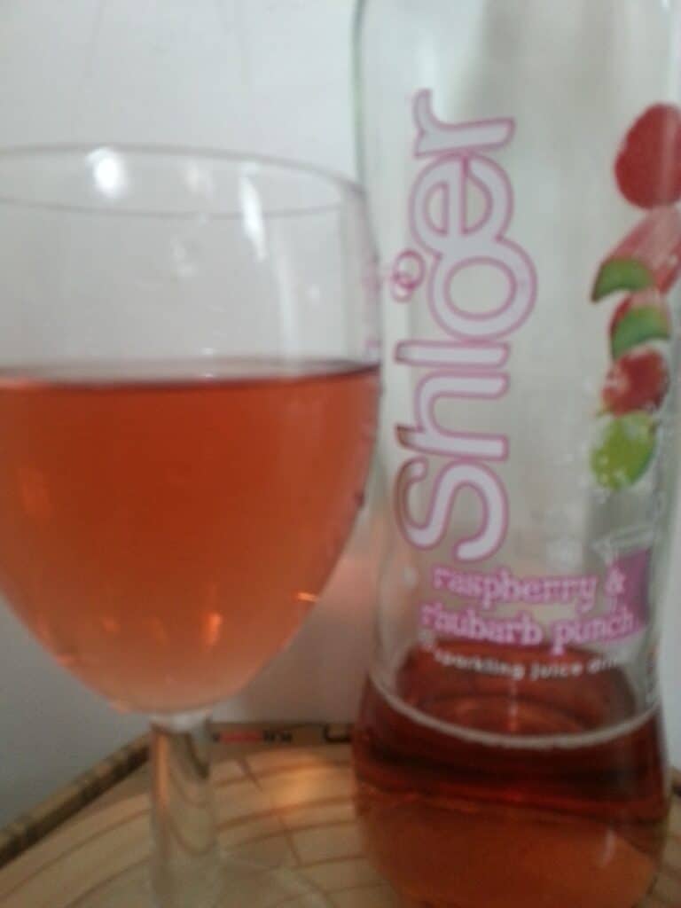 A glass of pink sparkling juice sits next to a bottle labelled "Shloer raspberry & rhubarb punch." The bottle has images of raspberries and rhubarb on the label, and both are on a light surface with a plain background.