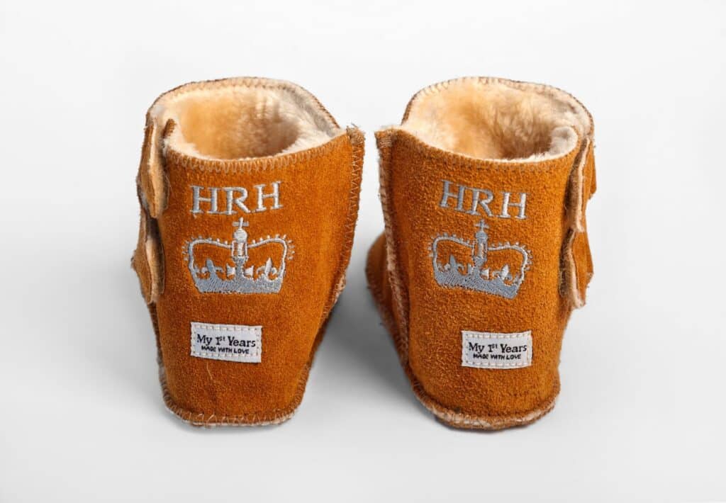 A pair of tan suede baby bootees with soft beige lining. Each bootee is embroidered with “HRH” above a silver crown and has a patch reading “My 1st Years: www.my1styears.com” on the heel. The shoes are shown from the back on a white background.