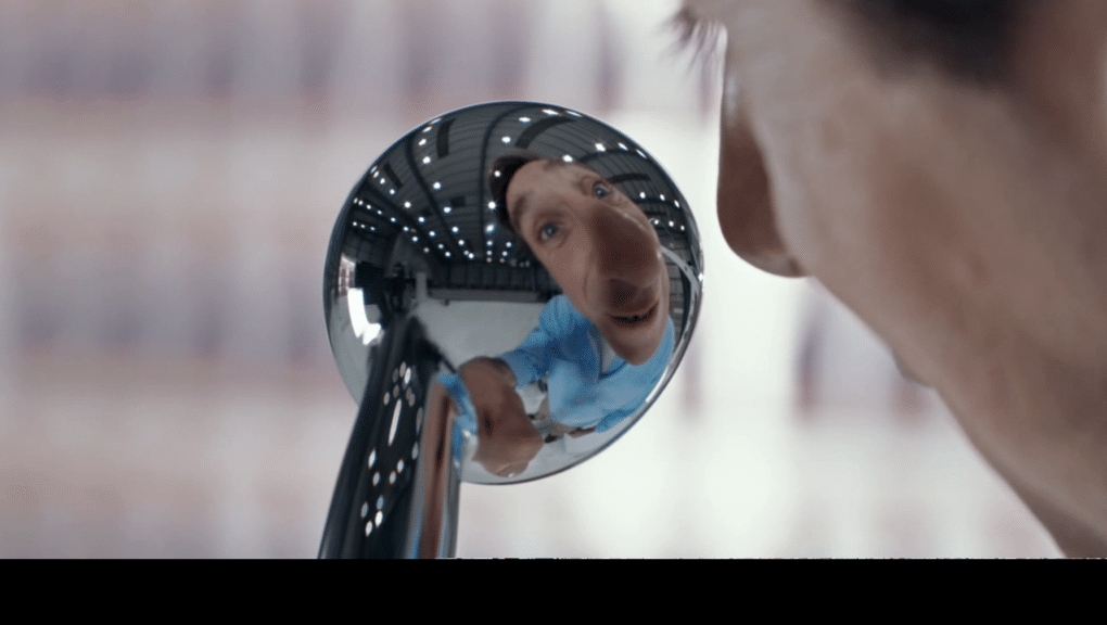 A person's face is distorted in the reflection of a round, convex mirror. The individual wears a light blue shirt and leans forward, with their features exaggerated by the curved surface. The background appears blurred, focusing on the curious expression.