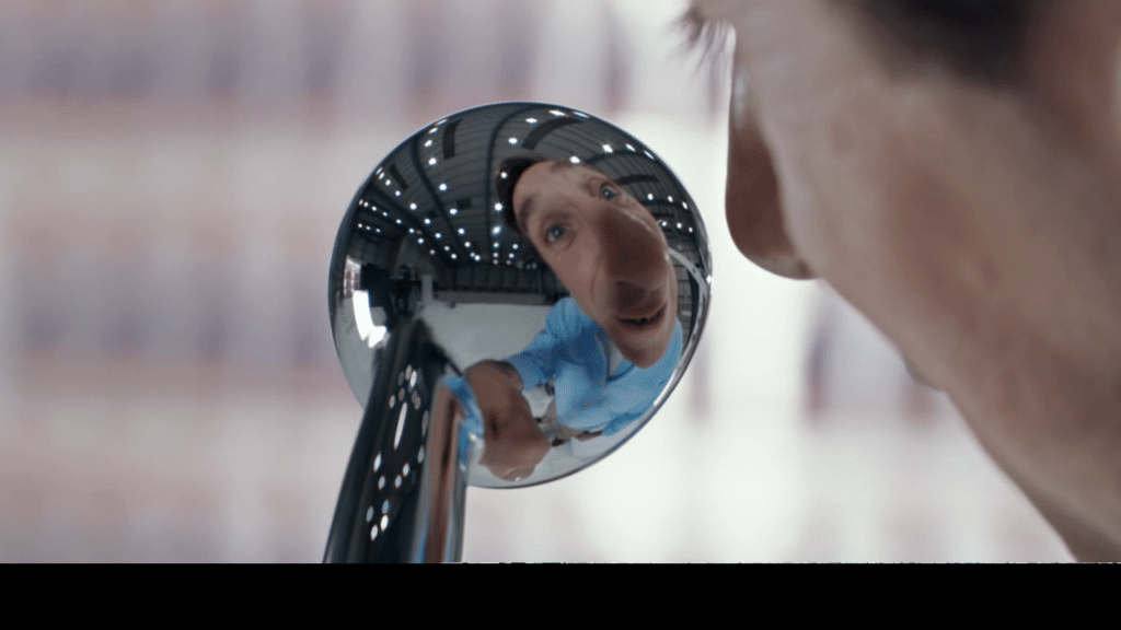 A man's distorted face and upper body are reflected in a small convex mirror, creating a fisheye effect. He is indoors, wearing a light blue shirt, with a blurred background featuring large windows and structural beams.