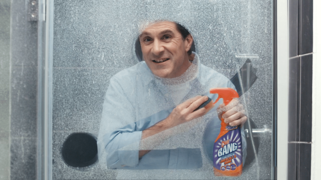 A man in a light blue shirt smiles whilst holding a bottle of Cillit Bang cleaner inside a shower with a frosted, soap-stained glass door. The cleaning product is in his right hand, and his left hand is poised as if about to clean the glass.