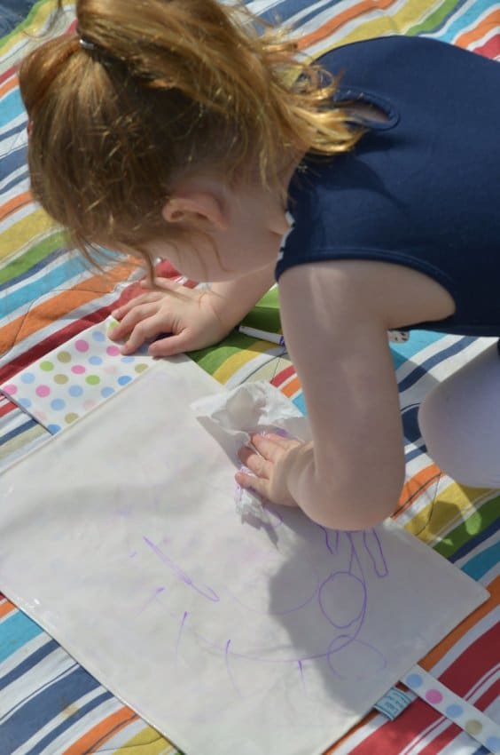A young child with light brown hair in a ponytail, wearing a navy sleeveless top, draws with a purple felt-tip pen on a white sheet of paper while kneeling on a colourful striped blanket outdoors. Her hand holds a tissue, dabbing the paper near scattered dot stickers.