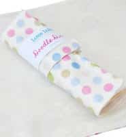 A rolled-up white baby blanket with pastel polka dots in pink, blue, green, and beige, wrapped with a white label that reads “Doodle&Co.” The blanket is placed on top of a plain cream-coloured surface.