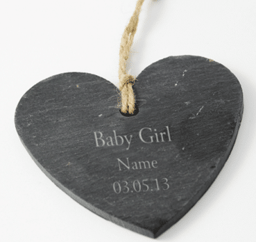 A dark, heart-shaped slate ornament with a hole at the top, through which a piece of string is threaded for hanging. The front is engraved with the words: “Baby Girl Name 03.05.13” in light-coloured text.
