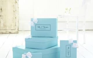 Four light blue gift boxes labelled "My 1st Years" are stacked on a white wooden floor. Each box has a white ribbon bow. There is an old distressed white door, a white chair, and bright natural light in the airy room.