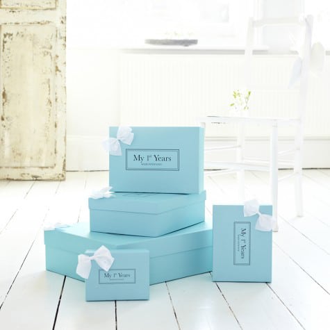 Four light blue gift boxes labelled "My 1st Years" are stacked on a white wooden floor. Each box has a white ribbon bow. There is an old distressed white door, a white chair, and bright natural light in the airy room.