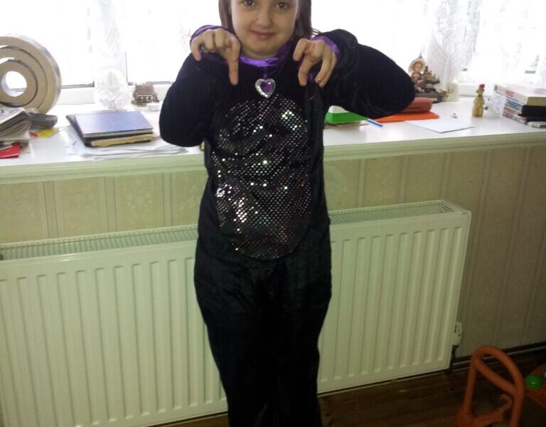 A child stands indoors dressed in a black cat costume with sequin details, cat ears headband, and a purple heart necklace. The child is making claw-like gestures with both hands. Behind them is a windowsill with books and ornaments.
