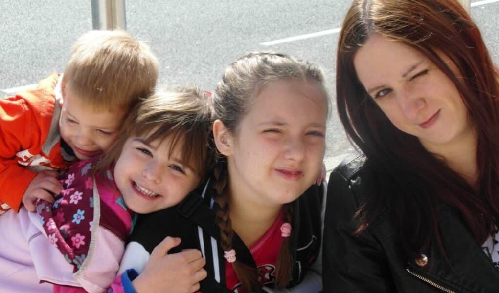 Four children and a woman sit close together outdoors. Two girls in the centre smile, one hugging a younger child in orange. The woman on the right has long brown hair and winks at the camera. The group appears happy and playful in bright daylight.