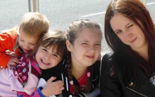 Four children and a woman sit close together outdoors. Two girls in the centre smile, one hugging a younger child in orange. The woman on the right has long brown hair and winks at the camera. The group appears happy and playful in bright daylight.