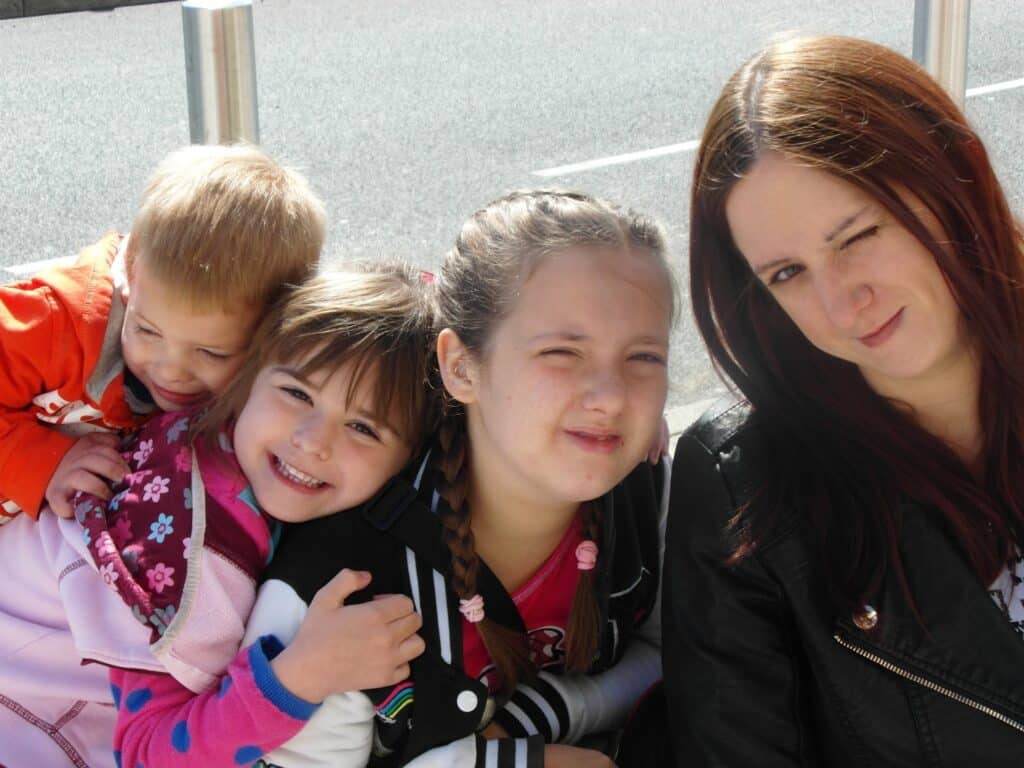 A woman with long brown hair winks at the camera, sitting beside three smiling children. Two girls with plaits and one boy in an orange jacket are close together, all outdoors on a sunny day. A street and silver poles are visible in the background.