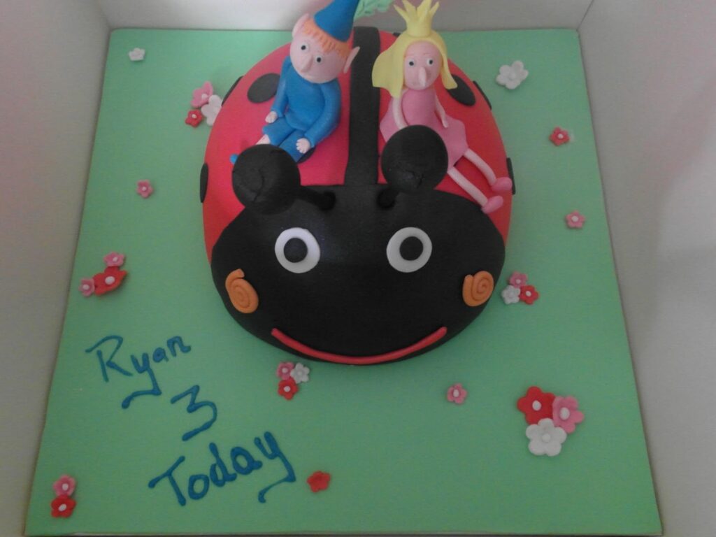A round ladybird-themed cake with large black eyes, antennae, and red wings topped with two fondant figures—a boy in blue with a cone hat and a girl in pink with a crown. “Ryan 3 Today” is written in blue on a green board decorated with small flowers.