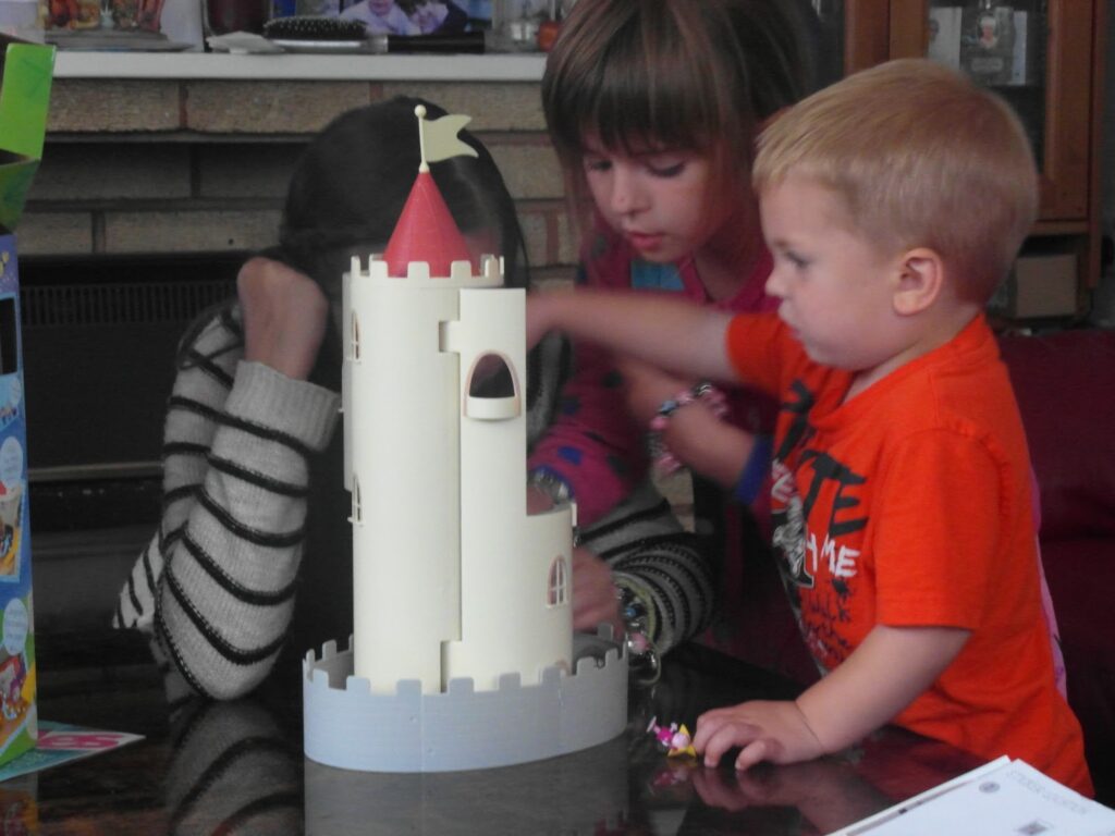 Three children play with a toy castle on a table indoors. One child in a striped jumper covers their face, while a girl with brown hair and a boy in an orange shirt focus on placing toy figures around the castle. A fireplace and shelves are in the background.