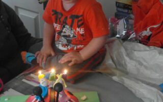 A young child in an orange T-shirt sits on a bed, smiling at a round ladybird-shaped birthday cake with lit candles. The cake is decorated with small figurines and pink flowers, and "Ryan 3rd Birthday" is written in blue icing on the cake board.