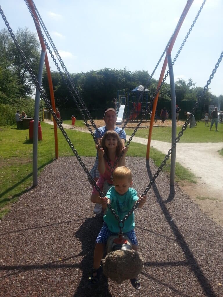 A woman and two young children sit together on a playground swing set. The boy in front holds the swing's chains and stands on the swing, while the girl sits behind him. The woman stands at the back, smiling, with a sunny park and play area in the background.