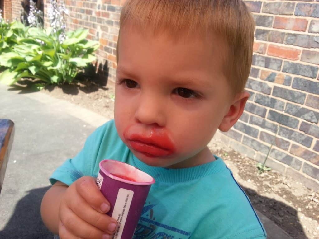 A young boy with short blonde hair is holding a purple ice cream tub, his mouth and lips smeared with red ice cream. He looks thoughtful or slightly pensive. Sunlight and outdoor plants are visible, with a brick wall in the background.