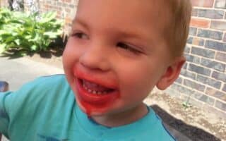 A young child with short blond hair smiles outdoors, wearing a light blue shirt. Red ice lolly stains surround his mouth, suggesting he just ate a treat. In the background, there is a brick wall, green plants, and sunlight illuminating the scene.