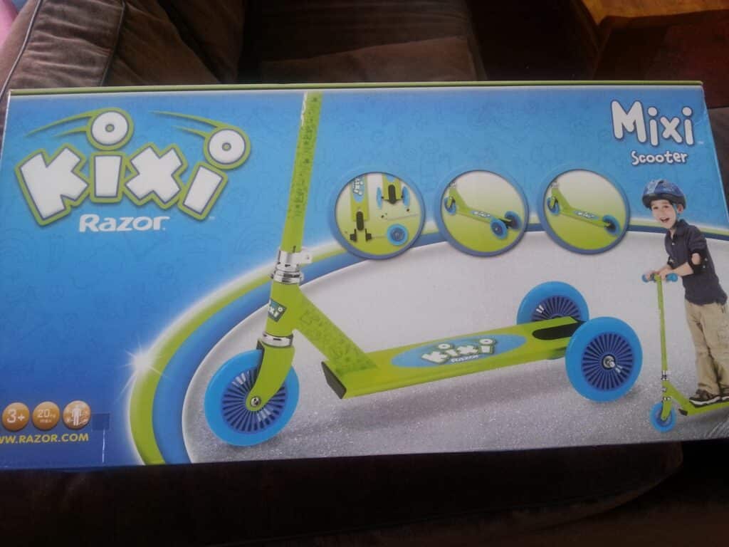 The image shows a box for a green and blue Kixi Razor Mixi Scooter. The packaging includes close-up images of the scooter’s wheels and handlebar, and a smiling child wearing a helmet stands next to the assembled scooter on the right.