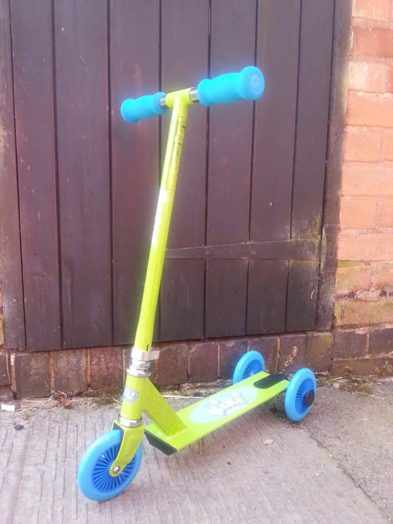 A bright green three-wheeled scooter with blue handles and wheels leans against a dark wooden door. The scooter has one front wheel and two rear wheels, and is standing on a concrete surface with part of a brick wall visible on the right.