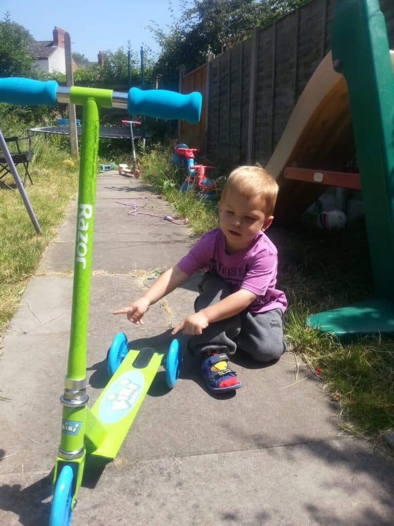A young child sits on a garden path, pointing at a bright green Razor scooter. The child wears a purple shirt, grey trousers, and colourful shoes. There is grass, a slide, and scattered toys in the background on a sunny day.