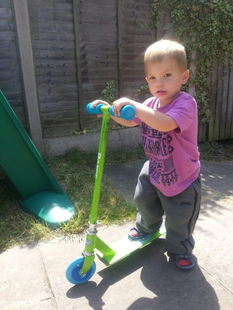 A young child with short blond hair, wearing a purple t-shirt, grey trousers, and blue sandals, stands outdoors on a green scooter. The child grips the blue handlebars and looks at the camera. There is grass, a slide, and a wooden fence in the background.