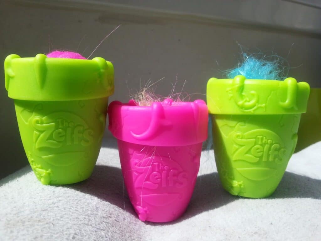 Three small plastic pots, two green and one pink, labelled “The Zelfs.” Each pot contains colourful fuzzy hair—pink, blue, and multicolour—sticking out of the tops. The pots are displayed on a light surface, with sunlight casting soft shadows.