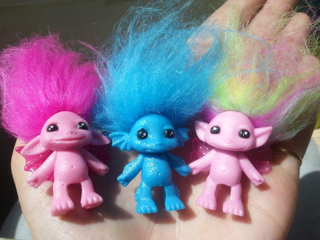 A hand holds three small troll dolls with big eyes and bright, fluffy hair. The dolls are pink with pink hair, blue with blue hair, and pink with rainbow hair. All have pointy ears and glittery bodies, standing together in the sunlight.