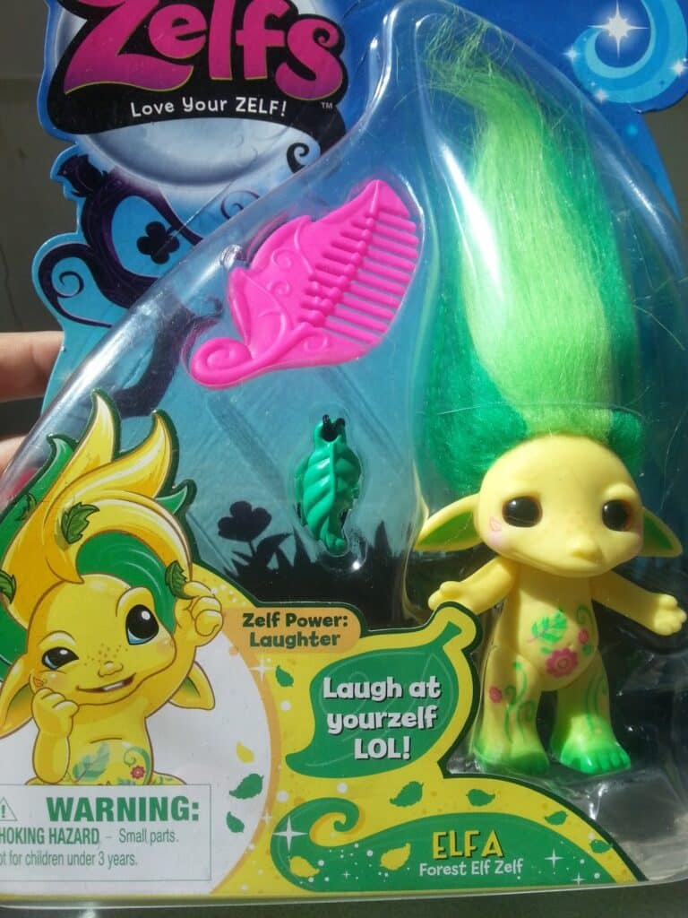 A toy package featuring "Elfa," a yellow Zelf doll with green leafy hair, leafy ears, and flower markings. Included are a pink comb and green hair accessory. Cartoon Elfa illustration, warning label, and the text “Laugh at yourself LOL!” are also visible.