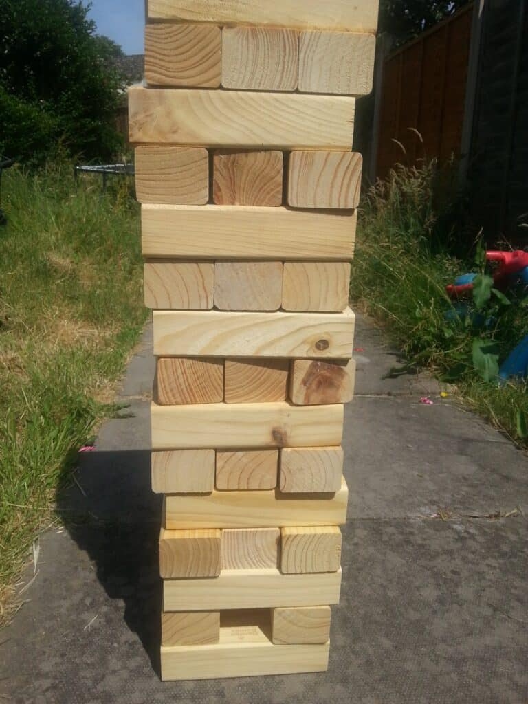 A large outdoor Jenga tower made of natural wooden blocks is stacked on a concrete path surrounded by grass. The tower consists of alternating layers of three blocks each, with some grass and garden items visible in the background.