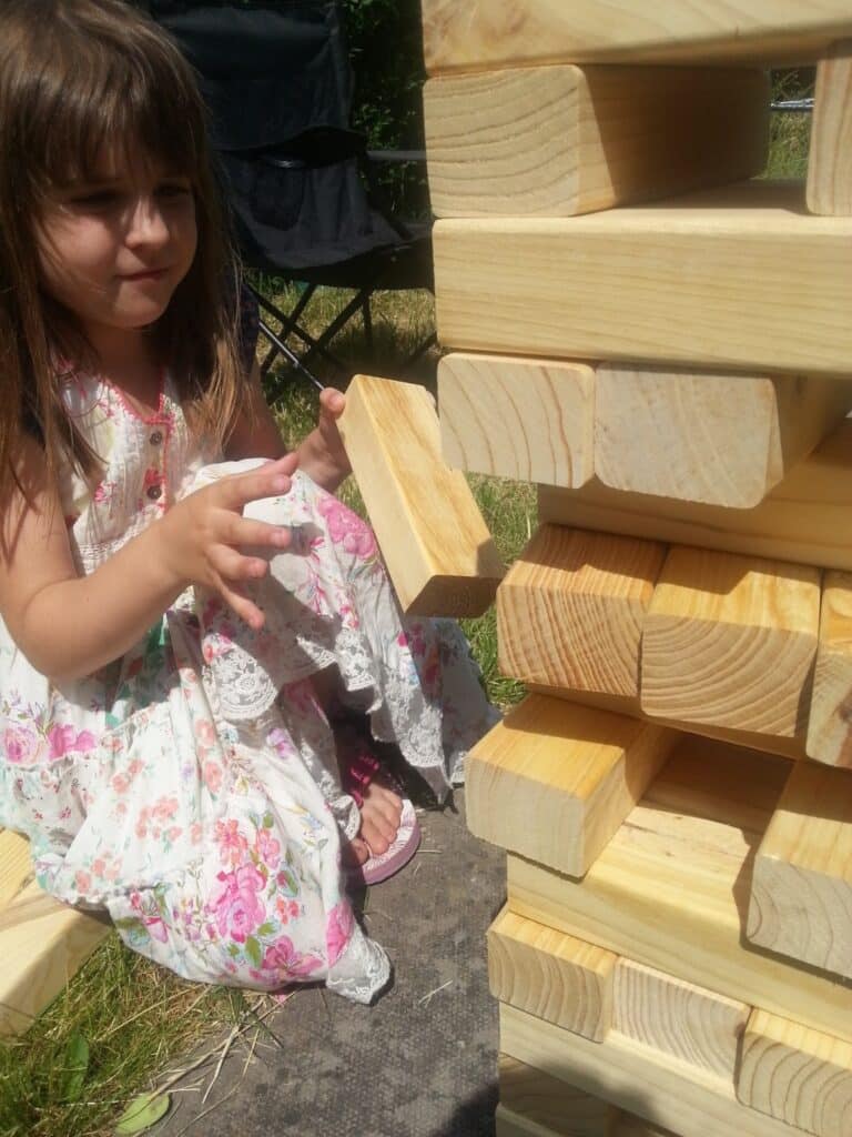 A young girl in a floral dress kneels beside a large outdoor wooden Jenga tower, carefully pulling out a block with both hands. Sunlight illuminates her hair and the wooden blocks, and green grass is visible in the background.