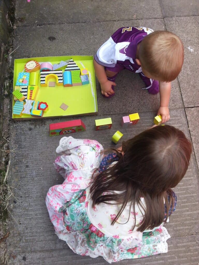 Two young children play outside on pavement with colourful wooden blocks and a yellow playset. The boy, wearing a purple and white shirt, is kneeling, and the girl, in a floral dress, is sitting as they build with the blocks together.