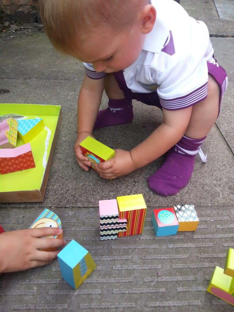 A young child in a white and purple outfit and purple socks is crouched on the ground, playing with colourful patterned wooden blocks. The child is arranging and stacking the blocks on a textured outdoor surface. Another child's hands are also visible, helping.