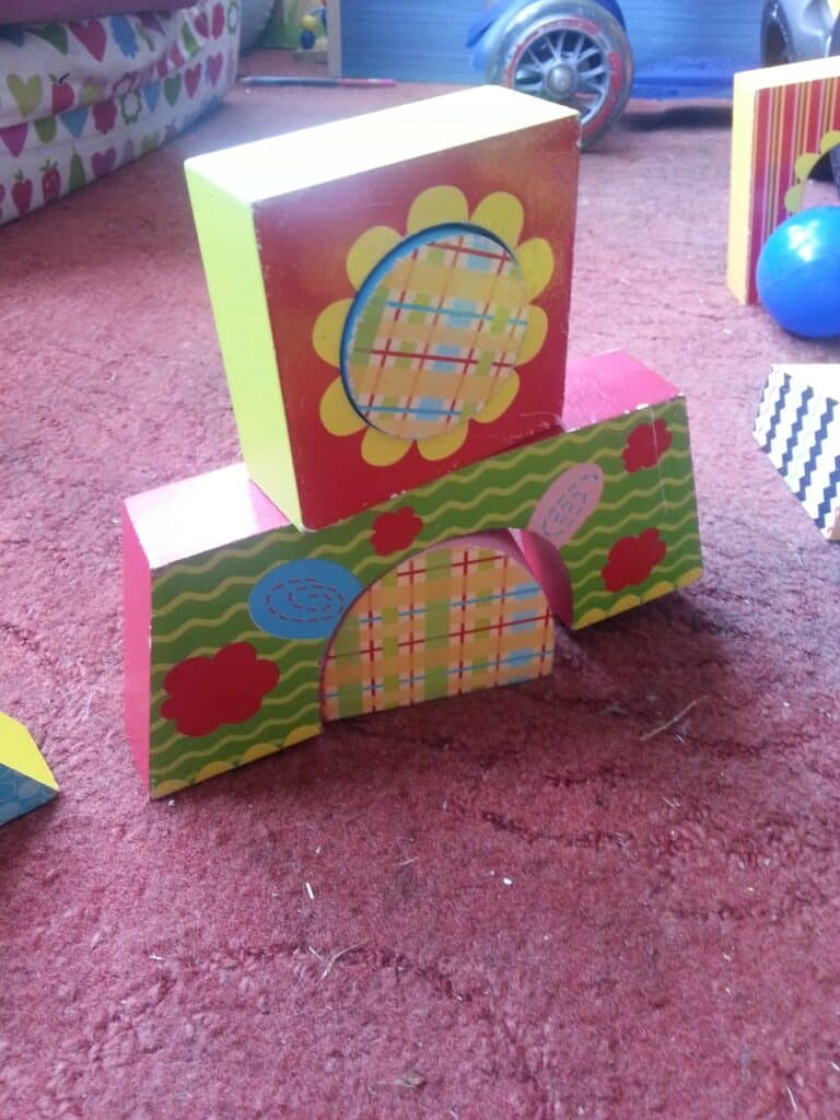 Three colourful wooden toy blocks are stacked on a red carpet. The bottom two red blocks are decorated with flowers, wavy green stripes, and a multicoloured arch. A yellow block with a flower and plaid pattern sits on top. Toys and a blue ball are in the background.