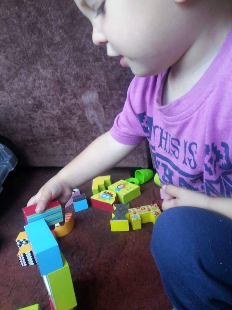 A young child wearing a purple shirt and blue trousers sits on a carpet, focused on stacking colourful wooden blocks. The child's hand is reaching to place a block, and various patterned blocks are scattered nearby. The background is a brown cushion.