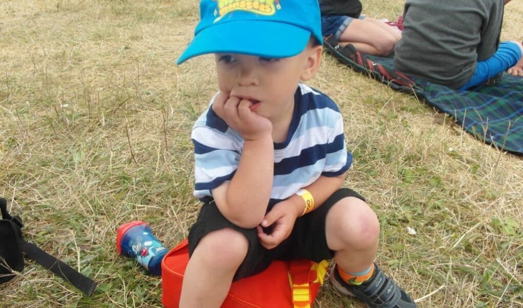 A young child wearing a blue "Moshi Monsters" cap, striped shirt, and black shorts sits on an orange box on dry grass, with one hand in his mouth. People sit on blankets in the background at an outdoor event.