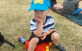 A young child wearing a blue "Moshi Monsters" cap, striped shirt, and black shorts sits on an orange box on dry grass, with one hand in his mouth. People sit on blankets in the background at an outdoor event.