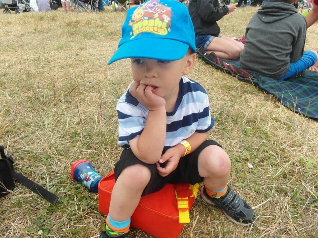 A young child wearing a blue "Moshi Monsters" cap, striped shirt, and black shorts sits on a red lunchbox, with one hand in their mouth. The child is outdoors on dry grass among other people sitting on blankets.