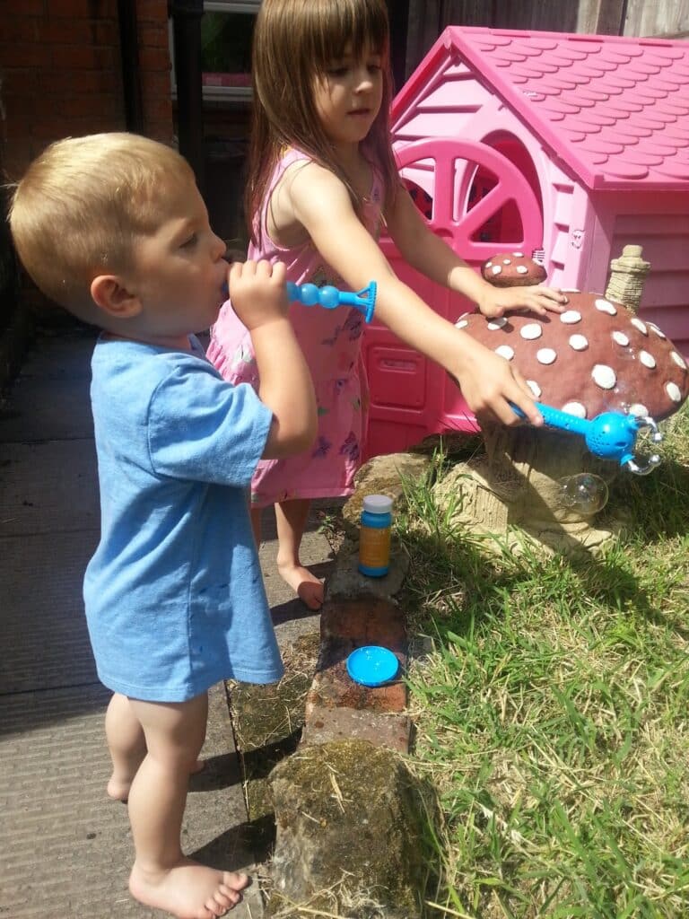 A young boy in a blue shirt and a girl in a floral dress blow bubbles outside by a large, decorative toadstool. Behind them is a bright pink toy house. Sunlight casts shadows, and both children appear focused on the bubble activity.