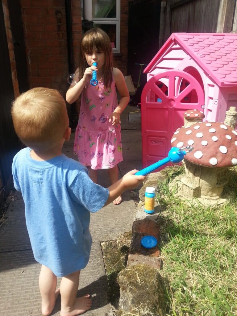 A girl in a pink dress and a boy in a blue shirt blow bubbles outside near a pink playhouse and a large mushroom-shaped ornament. The sun is shining, and bubble solution bottles are visible on a small brick structure.