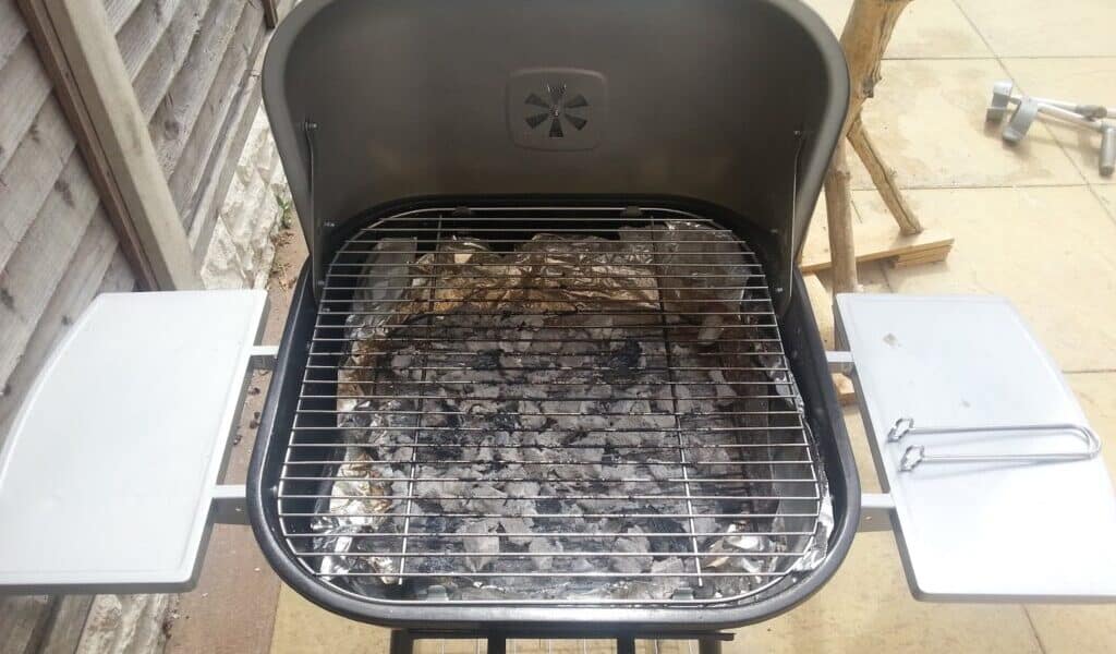 A charcoal barbecue stands on a patio, its lid open to reveal grey ash and scattered charcoal beneath a metal grid. Two metal side shelves and a barbecue tool are attached, with a wooden fence and tree branches visible in the background.