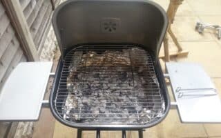A charcoal barbecue stands on a patio, its lid open to reveal grey ash and scattered charcoal beneath a metal grid. Two metal side shelves and a barbecue tool are attached, with a wooden fence and tree branches visible in the background.