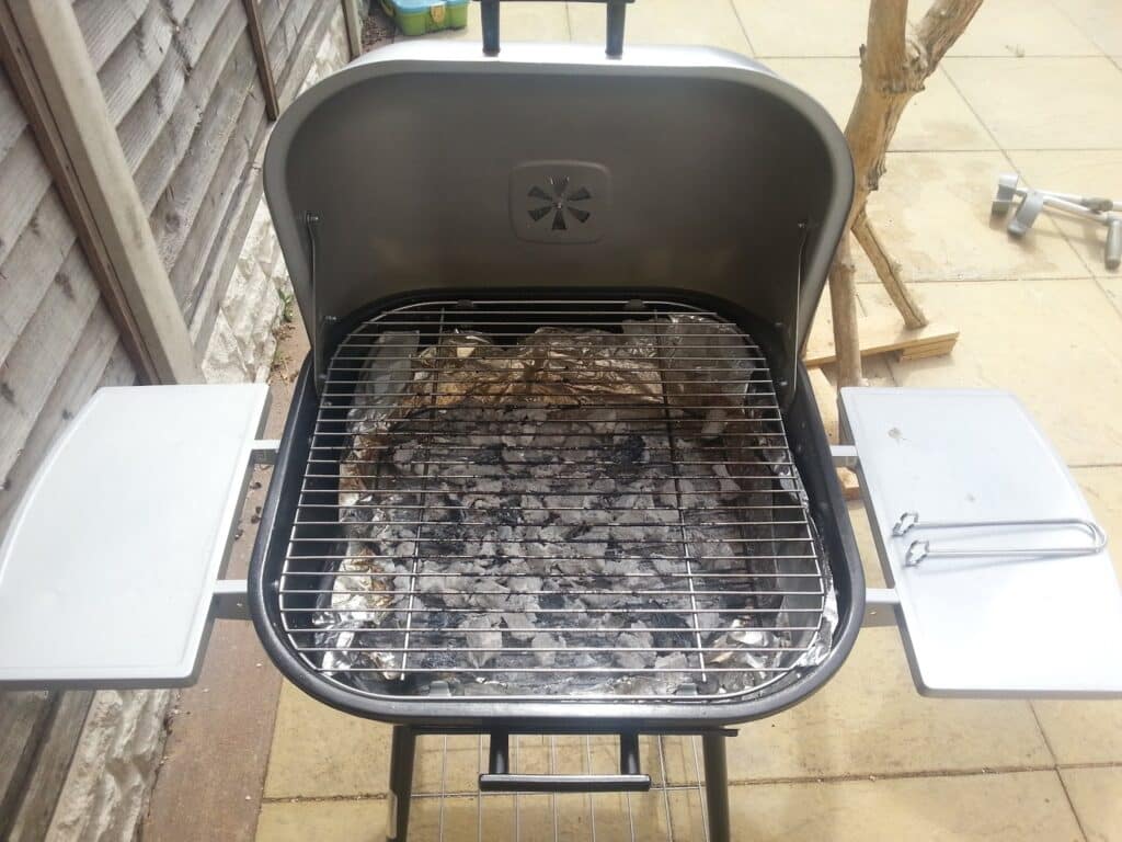 A charcoal barbecue grill with a partially open lid, two side shelves, and a metal grate over white ash-covered coals. The grill is on a tiled patio near a wooden fence and a tree branch, with outdoor items in the background.