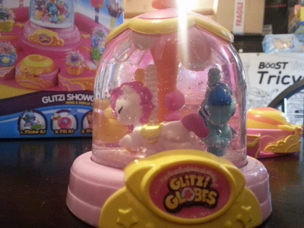 A close-up of a Glitzi Globes toy featuring small animal figurines inside a pink, dome-shaped case with glitter. The dome sits on a pastel yellow and pink base labelled "Glitzi Globes." The toy’s packaging is visible in the blurred background.