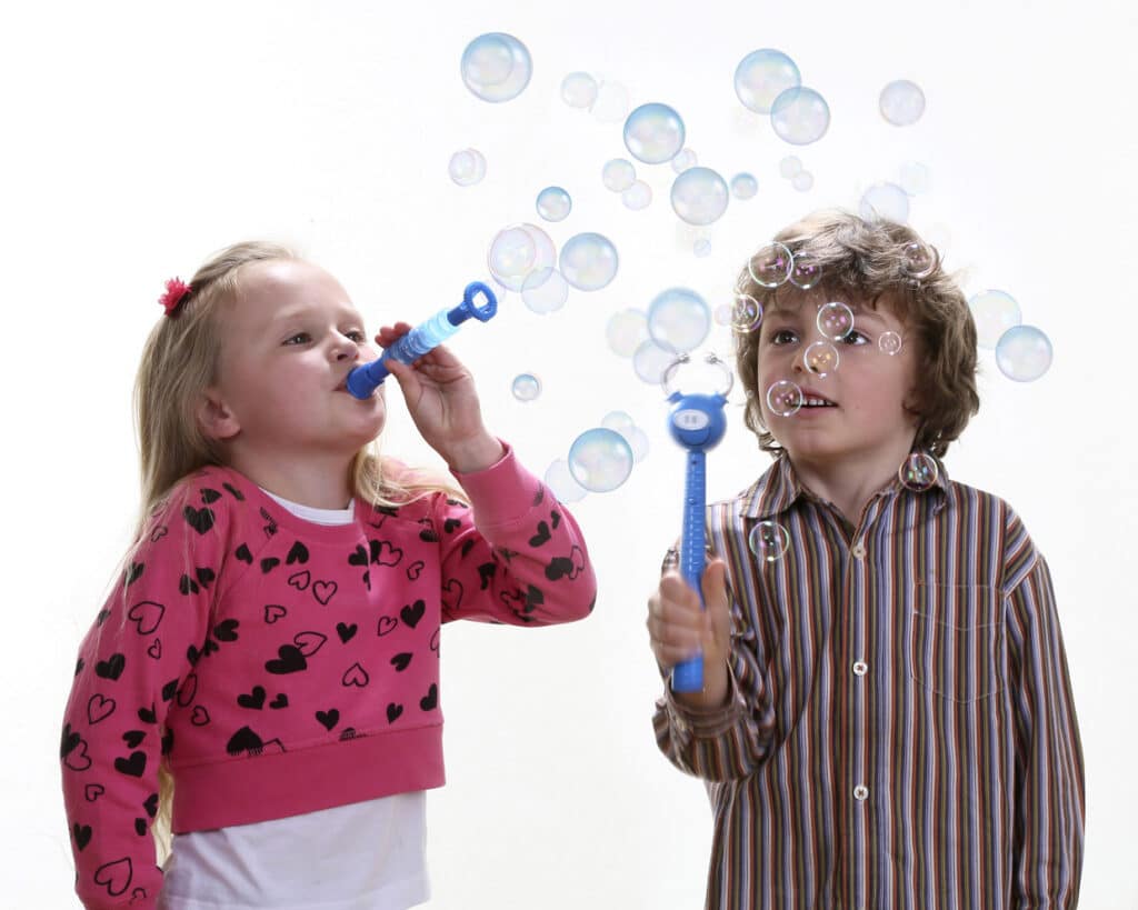 Two children stand side by side against a white background, blowing bubbles. The girl on the left wears a pink jumper with black hearts and blows bubbles with a blue wand. The boy on the right wears a striped shirt and smiles whilst holding a bubble wand. Bubbles float around them.
