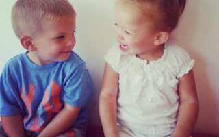 A young boy in a blue shirt and grey trousers sits next to a young girl in a white outfit with a ponytail. Both children are sitting against a plain light wall, smiling and looking happily at each other.